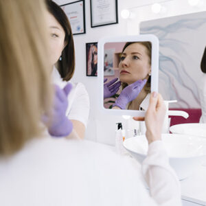 Reflection of calm female client sitting on couch in modern light salon while looking away during beauty procedure with mirror