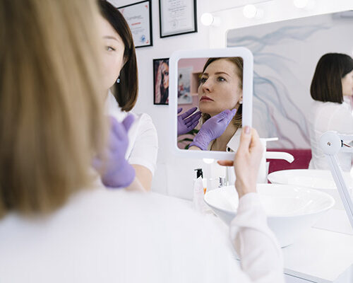 Reflection of calm female client sitting on couch in modern light salon while looking away during beauty procedure with mirror